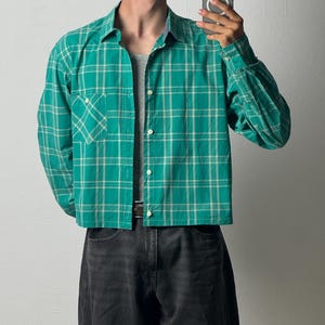 Può includere: Una camicia a maniche lunghe abbottonata con un motivo a quadri verde brillante e bianco. La camicia presenta un colletto classico, una tasca singola sul petto sul lato sinistro e un orlo dritto. È indossata sopra una canotta grigia e jeans neri.