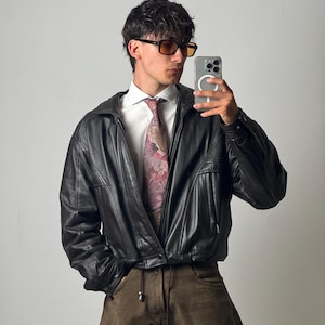 Könnte beinhalten: Eine schwarze Lederjacke über einem weißen Hemd mit Kragen und einer gemusterten rosa Krawatte, kombiniert mit einer braunen Hose. Das Outfit umfasst eine rechteckige Sonnenbrille mit bernsteinfarbenen Gläsern und ein Smartphone.
