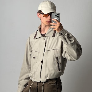 Può includere: Una giacca beige con colletto e finiture marrone scuro, dotata di chiusura frontale con bottoni, maniche lunghe con polsini abbottonati e una tasca con cerniera sul lato sinistro. È abbinata a un berretto da baseball beige e pantaloni marrone scuro.