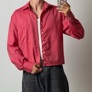 Può includere: Una camicia a maniche lunghe abbottonata di colore rosa scuro tinta unita con colletto classico, una tasca sul petto e bottoni bianchi. La camicia è indossata sopra una canotta bianca a coste e abbinata a jeans in denim grigio scuro.