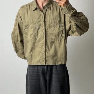 Peut inclure: Une chemise à manches longues boutonnée de couleur vert olive avec un col classique, deux poches de poitrine à boutons et une petite étiquette rectangulaire sur la poche droite. La chemise a un ourlet droit et est portée avec un jean gris foncé.