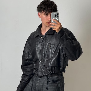 Può includere: Una giacca in pelle nera con silhouette corta, colletto a punta e chiusura frontale con zip. La giacca presenta una tasca sul petto con cerniera, maniche lunghe e un dettaglio con coulisse sull'orlo, abbinata a jeans in denim grigio scuro.