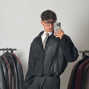 May include: A black leather jacket with a pointed collar, front button closure, and zippered side pockets. The jacket is worn over a white collared shirt and a patterned tie, paired with black trousers. The jacket features a cropped, boxy fit.