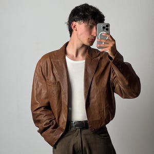 Könnte beinhalten: Ein brauner Lederblazer mit steigendem Revers, einem Knopfverschluss und langen Ärmeln. Der Blazer wird über einem weißen gerippten Tanktop und einer dunkelolivgrünen Hose mit einem schwarzen Ledergürtel mit silberner Metallschnalle getragen.