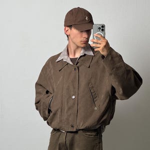 Könnte beinhalten: Eine braune Baseballkappe mit kleinem gesticktem Logo, eine braune Jacke mit Knopfleiste und Seitentaschen, ein helles kariertes Hemd mit Kragen und eine braune Hose. Das Outfit wird mit einem in der Hand gehaltenen Smartphone getragen.