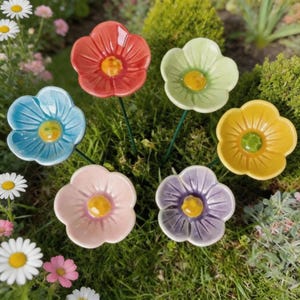 May include: A set of six ceramic flower garden stakes in red, light green, blue, yellow, pink, and purple. Each flower features five rounded petals and a raised yellow centre, attached to a thin green metal stem for placement in the ground.