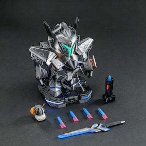 Könnte beinhalten: Eine metallisch silberne und schwarze Roboter-Actionfigur mit türkisfarbenen Visierakzenten, die auf einem Sockel mit blauen Lichtern steht. Enthalten sind eine kleine vogelartige Figur, vier blaue und orangefarbene Projektile, ein schwarzer Werfer und ein blau-silbernes Schwert.