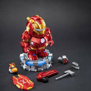 Puede incluir: Una figura estilizada de Iron Man con armadura roja y dorada sobre una base circular iluminada. El set incluye un pequeño casco con forma de pato, un escudo rojo, un bláster rojo, un martillo plateado y dos pequeños accesorios mecánicos.