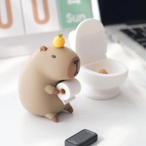 May include: A tan capybara figurine with a small yellow fruit on its head, holding a miniature roll of white toilet paper. Next to it is a small white toilet figurine with a tiny brown object inside the bowl and a black rectangular base nearby.