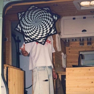 Könnte beinhalten: Ein sternförmiges Kissen mit einem schwarz-weißen karierten Spiralmuster. Das Kissen wird vor einer Person gehalten, die ein helles T-Shirt mit rotem Text und eine hellblaue Jeans mit einem Riss am linken Oberschenkel trägt.