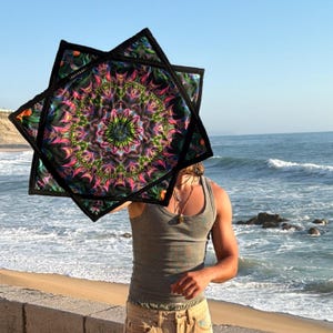 Puede incluir: Una pieza de tela cuadrada con un patrón de mandala simétrico y vibrante en tonos rosa neón, verde y morado sobre un fondo negro. La tela tiene un borde grueso cosido en negro y se sostiene para mostrar el diseño intrincado.