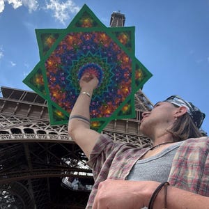 Puede incluir: Un paraguas en forma de estrella con un vibrante patrón de mandala caleidoscópico en tonos azul, morado, verde y naranja, con un borde verde sólido, sostenido contra un cielo azul claro con la Torre Eiffel visible al fondo.