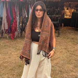 Könnte beinhalten: Ein rechteckiger Schal mit einem detaillierten Paisley-Muster in Orange-, Blau- und Brauntönen mit schwarzem Fransenbesatz. Der Schal ist über Kopf und Schultern drapiert und wird mit einem schwarzen bauchfreien Top und einer cremefarbenen weiten Hose kombiniert.