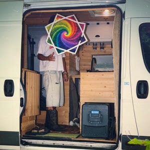 Può includere: Una persona si trova all'interno di un camper indossando una t-shirt bianca, pantaloncini di jeans chiari e stivali da cowboy neri. Una grande decorazione a forma di stella con un vivace motivo a spirale tie-dye arcobaleno nasconde il volto.