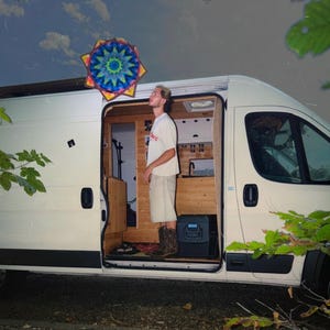 Könnte beinhalten: Ein weißer Campingbus mit offener Seitentür, die einen hölzernen Innenraum zeigt, eine Person im Inneren mit weißem T-Shirt und heller Shorts sowie eine bunte, sternförmige Mandala-Dekoration, die über dem Eingang des Vans hängt.