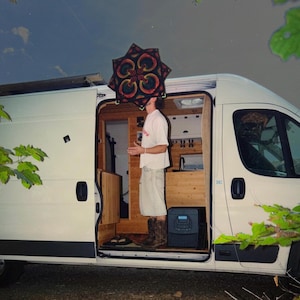 Pode incluir: Uma carrinha camper branca com a porta lateral aberta revelando um interior de madeira. Uma pessoa está de pé no interior vestindo uma t-shirt branca, calções claros e botas castanhas, segurando uma peça de tecido em forma de estrela com um padrão preto, vermelho e verde.