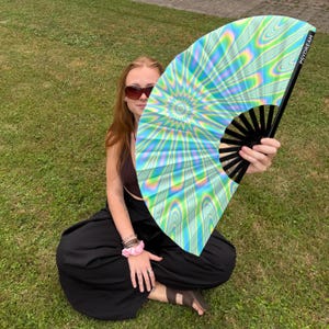 May include: A large handheld folding fan featuring a vibrant holographic pattern with swirling blue, green, and yellow hues. The fan has black plastic ribs and the word PSYDREAM printed in white text along the outer edge of the frame.
