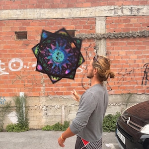 Puede incluir: Un objeto en forma de estrella de múltiples puntas con un diseño circular psicodélico vibrante y un motivo solar central en tonos azules, morados y verdes, sostenido en el aire frente a un muro de ladrillo rojo desgastado con grafitis.