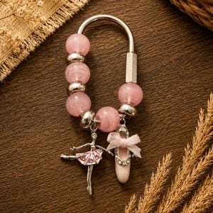 May include: A silver keychain with pink beads and charms. The charms include a ballerina and a ballet slipper with a pink bow. The background is a dark wood surface.