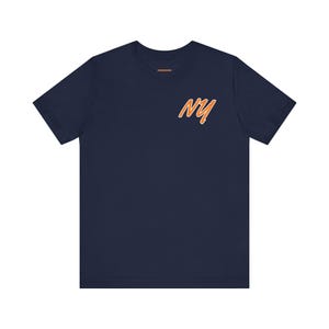 May include: A navy blue short-sleeve t-shirt featuring the letters NY in a stylized orange font with a white outline on the upper right chest area. The shirt has a crew neckline and a small orange brand label printed inside the collar area.