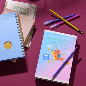 May include: A collection of notebooks and writing supplies on a purple surface. A blue spiral-bound notebook with a gold spiral is next to a notebook with the text "GREAT THINGS". A notebook with the text "Mon cahier d'écriture" is in the foreground.