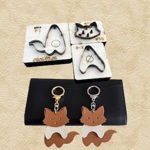 May include: A collection of leathercraft tools and fox-shaped keychains. The tools include metal dies for cutting leather, with the date "2026.05.26" visible. Two keychains feature a fox design in brown, white, and tan leather, attached to gold-colored keyrings.