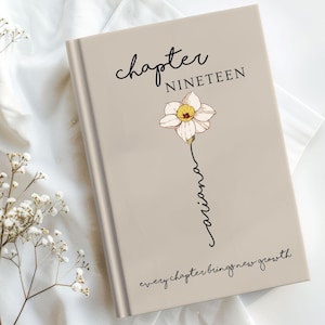May include: A beige hardcover book featuring the text "chapter NINETEEN" at the top, a central illustration of a white and yellow daffodil with the name "ariana" written in cursive as the stem, and "every chapter brings new growth" at the bottom.