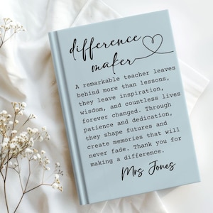 May include: A light blue notebook with the words "difference maker" and a heart symbol. The notebook has a quote about teachers and is signed "Mrs. Jones". The notebook is on a white surface with floral accents.