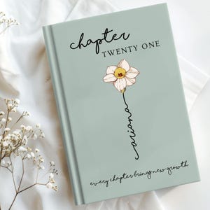 May include: A sage green hardcover book featuring the text Chapter TWENTY ONE at the top, a central illustration of a pale pink daffodil with the name Ariana written in script down the stem, and the phrase every chapter brings new growth below.