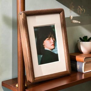 May include: A wooden picture frame displaying a portrait. The frame is crafted from dark wood with a substantial border. The portrait is set within a white mount. The frame rests on a wooden shelf alongside books and a small plant.