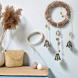 May include: A circular grapevine wreath with hanging brass bells and decorative charms. The wreath features three bells suspended by twine, along with metal charms. The background is a light blue wall with decorative items.