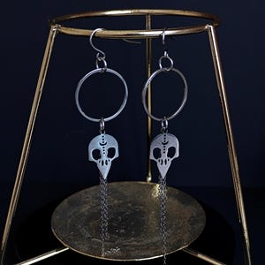 Peut inclure: Boucles d'oreilles argentées avec un motif de crâne. Chaque boucle d'oreille présente un pendentif en forme de crâne avec un détail de croissant de lune, suspendu à un cercle argenté. De fines chaînes pendent du bas des pendentifs en forme de crâne. Les boucles d'oreilles sont exposées sur un présentoir doré sur fond sombre.