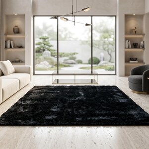 May include: A rectangular black area rug with a thick, shaggy pile texture. The rug is positioned centrally on a light-colored stone floor in a room featuring a cream-colored sofa, two dark gray rounded armchairs, and a glass coffee table.