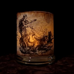 May include: A glass candle holder with a vintage illustration of a woman riding in a chariot pulled by wolves. The candle holder is lit and the light shines through the illustration.