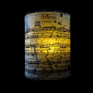 May include: A candle holder made from a vintage sheet music page. The sheet music is for "Arioso from Cantata No. 136". The candle is lit and glowing.