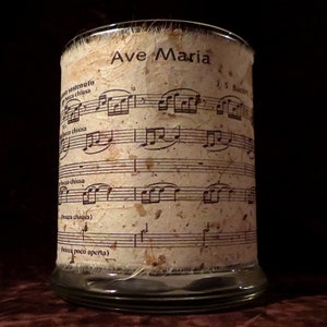 May include: A glass candle holder with a white paper label that has the words "Ave Maria" and musical notes printed on it. The label is slightly distressed and has a rustic look.