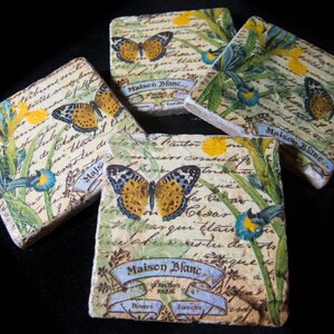 May include: Four square ceramic coasters with a vintage floral design featuring butterflies and handwritten text. The text includes the words "Maison Blanc" and "Diners Lanchs".