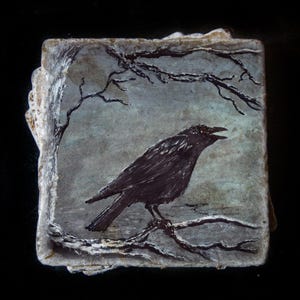 May include: A square stone coaster featuring a hand-painted black crow perched on a bare tree branch. The background is a muted, textured grey-blue sky with additional dark, leafless branches extending across the top of the square surface.