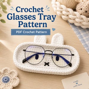 May include: A white oval crochet tray with two small ears, resembling a rabbit face, holding black-rimmed glasses. The text reads Crochet Glasses Tray Pattern and PDF Crochet Pattern. A small card says made with love and a crochet flower is nearby.
