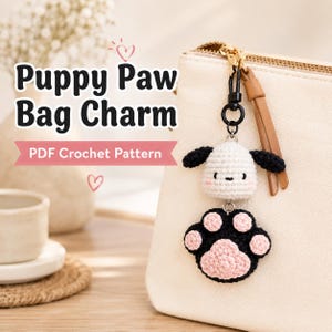 May include: A crochet bag charm featuring a small white puppy head with black ears and a black paw pad with pink accents. The charm hangs from a gold-toned metal clasp attached to a cream-colored bag. Text reads Puppy Paw Bag Charm PDF Crochet Pattern.