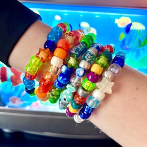 May include: A collection of multiple stacked bracelets made from colorful plastic pony beads in various shades of blue, green, yellow, orange, red, and white. The bracelets feature assorted shapes including round spheres, translucent stars, and a small fish charm.