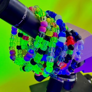 May include: A collection of multi-colored plastic pony bead bracelets featuring a vibrant mix of neon green, bright pink, deep blue, black, and translucent beads. The bracelets are stacked together and wrapped around a black cylindrical object.