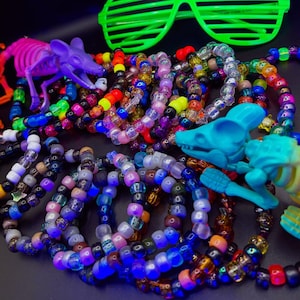 May include: A collection of colorful plastic pony bead bracelets arranged with a bright green pair of shutter shades and two plastic animal skeletons, one purple and one teal, featuring a variety of translucent and opaque beads.