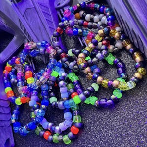 May include: A collection of circular bracelets made from multi-colored plastic pony beads. The beads feature a variety of finishes including translucent, opaque, metallic, and neon colors such as blue, purple, green, orange, and yellow.