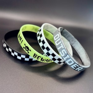 May include: Three silicone wristbands stacked together. One is black with a white checkerboard pattern. Another is neon yellow with a black skull icon and the text BEWARE. The third is white with a black checkerboard pattern and the word OUTSIDER.