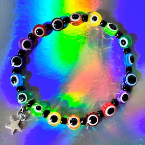Può includere: Un braccialetto circolare con perline multicolori a forma di occhio in tonalità rosso, arancione, giallo, verde, blu, viola e marrone, separate da piccole perline distanziatrici nere. Un ciondolo a forma di stella in metallo argentato pende.