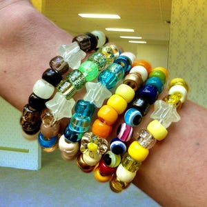 May include: A collection of multiple stacked bracelets made from colorful plastic pony beads in various shades of blue, yellow, green, orange, red, white, and black, featuring star-shaped beads, clear beads, and round beads with eye patterns.