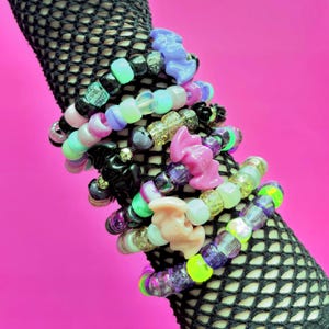 May include: A stack of colorful beaded bracelets featuring various plastic bat-shaped charms. The beads are a mix of translucent, opaque, glittery, and glow-in-the-dark finishes in shades of purple, pink, green, black, white, and blue.