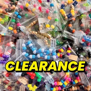 Könnte beinhalten: Eine Sammlung von bunten Pony-Perlen-Armbändern aus Kunststoff, die einzeln in durchsichtigen Plastiktüten verpackt sind. Die Armbänder weisen verschiedene Farbkombinationen und Muster auf, wobei einige Tüten beschriftet sind. Das Wort CLEARANCE ist in fettem gelben Text darübergelegt.