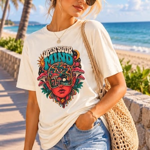 May include: An ivory t-shirt featuring a colorful graphic print with the text "OPEN YOUR MIND" above a stylized face, mushrooms, and celestial elements. The design includes shades of red, blue, and yellow, with a woven beige shoulder bag.